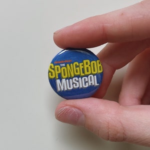 May include: A round, blue button featuring the text "THE SPONGEBOB MUSICAL" in yellow and white, with the Nickelodeon logo. The button is held between two fingers.