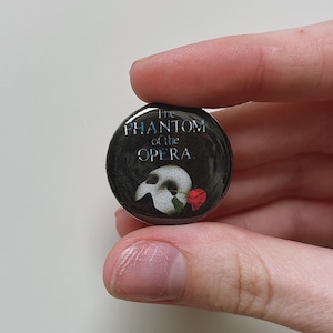 Puede incluir: Un botón redondo con el texto "The Phantom of the Opera" en blanco y azul. El diseño incluye una máscara blanca, una rosa roja y un fondo oscuro.