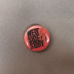 May include: A round, red button with the words "WEST SIDE STORY" in black, along with silhouettes of dancers and a ladder. The button is on a gray surface.