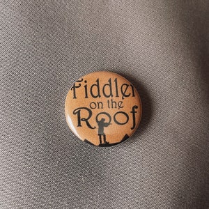 May include: A round, tan button with the words "Fiddler on the Roof" in black script. A small silhouette of a person playing a violin is below the text. The button is on a gray surface.