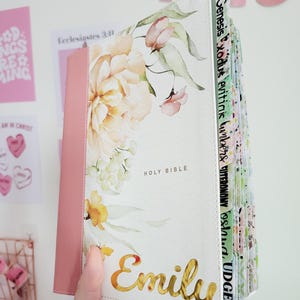 May include: A white Holy Bible with a floral design and the name "Emily" in gold lettering. The spine is pink. The pages have colorful tabs with book names. The words "Holy Bible" are printed on the cover.