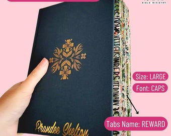 May include: A navy blue ESV Study Bible with gold floral accents and the name "Prandee Shelton" in gold lettering. The book has labeled tabs and the text "ESV Women's Study Bible". The size is large, with text in caps.