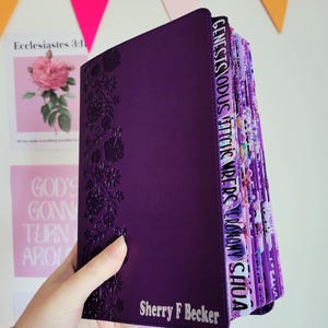 May include: A purple Bible with floral embossed detailing and labeled tabs. The cover has the name "Sherry F Becker" and the word "thanks" printed on it. The Bible is held in front of a white wall with decorative paper cutouts.