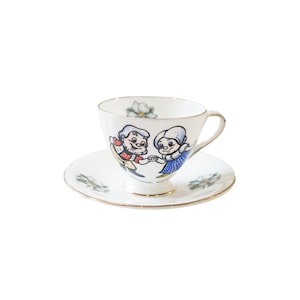 May include: A white teacup and saucer set with a gold rim. The cup and saucer feature a cartoon illustration of a couple dancing, surrounded by white flowers.