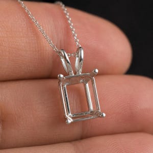 Pode incluir: Um colar com pingente de prata com uma moldura retangular. O pingente tem quatro garras em cada canto para segurar uma pedra preciosa. A corrente é delicada e prateada.