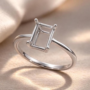 Pode incluir: Um anel de prata com uma cravação de corte esmeralda. O anel apresenta uma banda polida e uma cravação retangular com quatro garras em cada canto para segurar uma pedra preciosa. O anel é exibido sobre um fundo neutro.