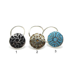 Può includere: Tre anelli da naso circolari con sottili cerchi in argento. Il primo presenta un intarsio nero con un motivo floreale argentato, il secondo un intarsio color oro metallizzato con un design geometrico e il terzo un intarsio turchese brillante.