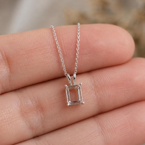 Pode incluir: Um delicado colar de prata com um pingente retangular. O pingente tem um design claro e aberto, com quatro pequenas garras em cada canto. A corrente é fina e parece ser de prata. O colar é exibido em um fundo macio e neutro.