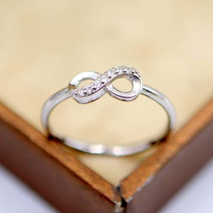 Pode incluir: Um anel prateado com um design de símbolo de infinito. O símbolo do infinito é adornado com pequenas pedras preciosas transparentes. O anel é apresentado sobre um fundo macio e neutro, possivelmente numa exposição de joias.