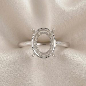 Pode incluir: Uma base de anel de prata com um centro oval. O anel tem quatro garras para segurar uma pedra preciosa. A banda é lisa e polida. O anel é exibido em um tecido creme.