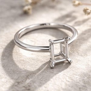 Op de afbeelding: Een zilveren ring met een vierkante zetting, ontworpen om een edelsteen te bevatten. De ringband is glad en gepolijst, en de zetting heeft vier klauwen. De ring wordt getoond op een lichtgekleurd oppervlak.