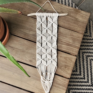 May include: A white macrame wall hanging with a geometric pattern. The hanging is made of cotton cord and has a wooden dowel at the top.