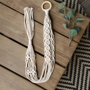 May include: A macrame plant hanger with a wooden ring at the top. The hanger is made of natural cotton cord and has a delicate, intricate pattern. The hanger is hanging on a wooden surface.