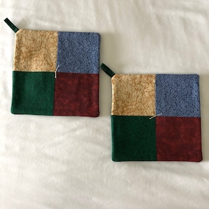 May include: Two quilted pot holders with a patchwork design. The pot holders are made of four squares in beige, blue, green, and burgundy.