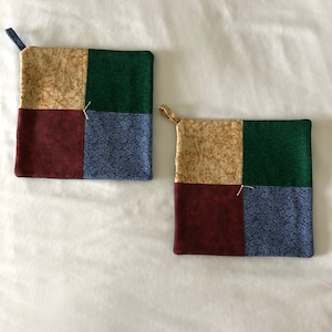 May include: Two square pot holders with a patchwork design. The pot holders are made of four squares of fabric in beige, green, burgundy, and blue.