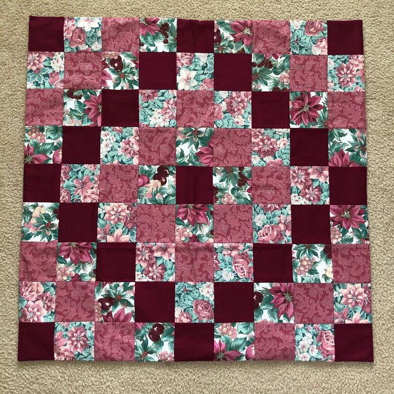 May include: A square patchwork quilt featuring a checkerboard pattern of solid deep burgundy squares alternating with squares displaying a floral print of poinsettias, holly berries, and green leaves on a white and muted red background.