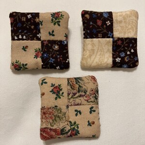 May include: Three square fabric coasters with a patchwork design. Each coaster is divided into four squares, with alternating floral and solid tan patterns. The floral patterns feature red, blue, and green flowers on a dark brown background.
