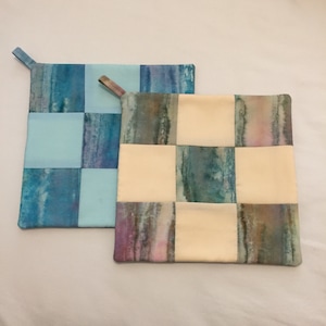 May include: Two quilted pot holders, one blue and white and one with a cream background and blue, green, and pink watercolor-like designs.