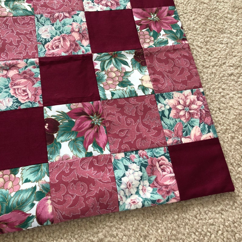 May include: A patchwork quilt featuring a repeating grid of solid deep burgundy squares alternating with squares displaying floral and botanical patterns in shades of burgundy, sage green, and white, creating a structured textile design.