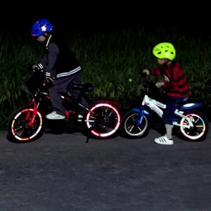 May include: Two children riding bicycles at night with reflective wheel rim stickers. The child on the left is wearing a blue helmet and the child on the right is wearing a yellow helmet.