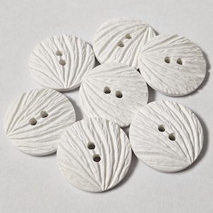 May include: Seven white, circular buttons with a textured, ridged pattern. Each button features two holes for sewing. The buttons are displayed on a white surface, highlighting their design. These buttons are ideal for sewing and craft projects, with a diameter of approximately 2.5 cm.