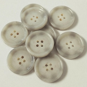 May include: A collection of eight round, light gray buttons with four holes each. The buttons have a marbled appearance with subtle variations in color, and are arranged in a loose pile.
