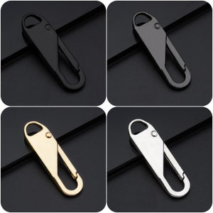 Könnte beinhalten: Vier Metall-Reißverschluss-Anhänger in Schwarz, Gunmetal, Gold und Silber. Jeder Anhänger hat eine Schlaufe oben und einen federbelasteten Verschluss. Die Anhänger werden vor einem dunklen Hintergrund gezeigt.