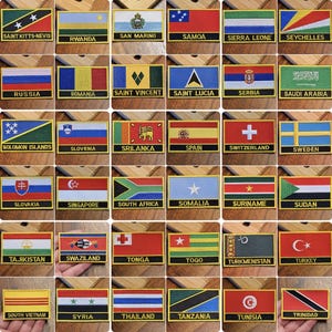 May include: A collection of 36 embroidered patches featuring flags from different countries. The patches are rectangular and have a gold border. Each patch has the name of the country printed below the flag.
