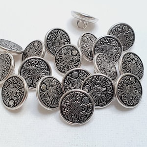 Puede incluir: Una colección de botones plateados con diseños en blanco y negro. Los botones presentan un patrón repetido de un escudo de armas con una corona y otros detalles.