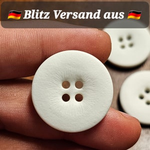 Könnte beinhalten: Vier weiße Knöpfe mit jeweils vier Löchern. Die Knöpfe sind glatt und haben eine leicht strukturierte Oberfläche.