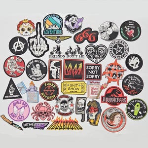 Könnte beinhalten: Eine Sammlung von aufgestickten Aufnähern mit verschiedenen Designs, darunter Totenköpfe, Mittelfinger und Texte wie "Adventure" und "Jurassic Park". Die Aufnäher weisen eine Mischung aus Farben und Stilen auf.