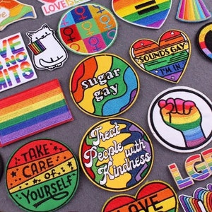 Op de afbeelding: Een assortiment strijkbare patches in regenboogkleuren met verschillende boodschappen, waaronder "Sugar Gay", "Treat People with Kindness", "Take Care of Yourself" en "Love is Love".