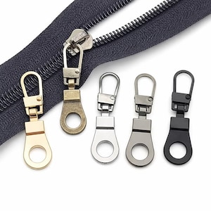 May include: Close-up of a dark gray zipper with a silver pull tab and five metal zipper pulls in gold, bronze, silver, gray, and black. Each pull has a ring at the bottom for easy attachment.