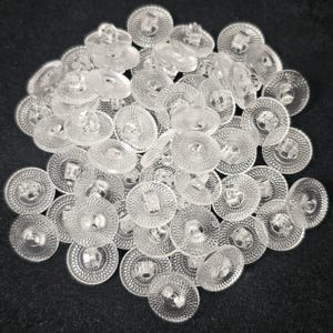 Puede incluir: Un montón de botones redondos y transparentes con un diseño de círculos concéntricos texturizados. Cada botón tiene un pequeño vástago rectangular y un centro ligeramente elevado. Los botones son de material transparente y están dispersos sobre una superficie oscura.