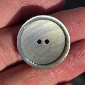 Puede incluir: Primer plano de un botón redondo nacarado con dos pequeños agujeros. El botón tiene un borde ligeramente elevado y un sutil patrón arremolinado. El botón es de color claro y neutro, y parece estar hecho de un material natural.
