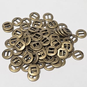 Pode incluir: Uma coleção de pequenas fivelas redondas de metal com cor de latão antigo. Cada fivela tem uma abertura quadrada ou retangular no centro e duas barras paralelas de um lado. As fivelas estão espalhadas sobre uma superfície branca.