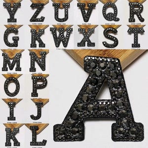 Könnte beinhalten: Eine Sammlung von schwarzen, mit Strasssteinen besetzten Buchstaben, jeweils auf einer Holzbasis montiert. Die Buchstaben variieren in der Größe, wobei der Buchstabe "A" prominent dargestellt wird. Ideal für personalisierte Bastelarbeiten und Verzierungen.