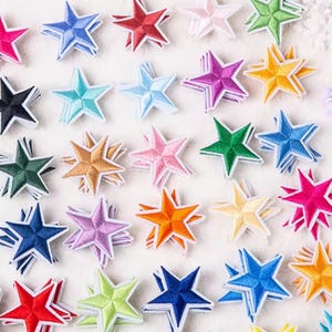 Puede incluir: Una colección de parches de estrellas bordadas en varios colores, incluyendo azul, rojo, rosa, verde y amarillo. Cada estrella tiene un diseño en capas con costuras blancas, creando un efecto 3D. Los parches están dispuestos sobre una superficie blanca.