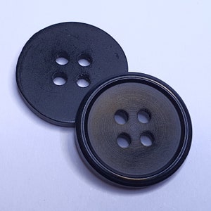 Puede incluir: Dos botones negros con cuatro agujeros cada uno. El botón de la derecha tiene una superficie ligeramente elevada y texturizada.