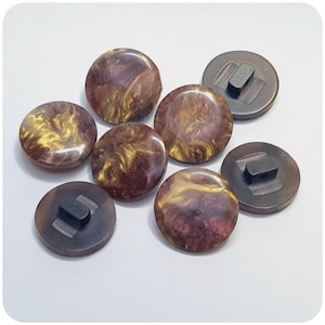 Op de afbeelding: Zeven bruine en gouden swirl-knoppen met zwarte plastic ruggen. De knoppen hebben een marmer effect met gouden en bruine swirls.
