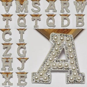 Könnte beinhalten: Eine Sammlung von Buchstaben, die mit Kunstperlen und Strasssteinen verziert sind. Die Buchstaben sind in verschiedenen Schriftarten und Größen gehalten, wobei der Buchstabe "A" im Vordergrund steht. Jeder Buchstabe ist etwa 2,4 bis 5,8 cm hoch.
