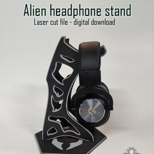 Könnte beinhalten: Ein schwarzer, lasergeschnittener Kopfhörerständer in Form eines Alienkopfes. Der Ständer hat eine sechseckige Basis und ein Design, das einem Alien-Gesicht ähnelt. Der Ständer hält einen schwarzen Kopfhörer mit einem silbernen Logo.