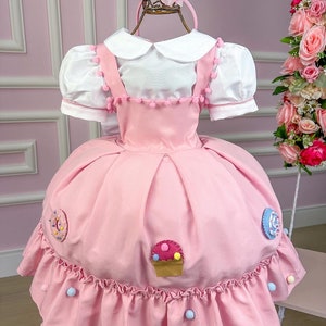 May include: A pink dress with a white Peter Pan collar and puff sleeves. The dress has a skirt with a ruffle trim and is decorated with felt cupcake, donut, and lollipop appliques. The dress is adorned with pink pom-poms.