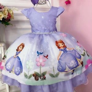 May include: A lavender princess dress with a white skirt featuring a cartoon princess design, flowers, and woodland creatures. The dress has short sleeves, a sparkly belt, and a matching bow headband.