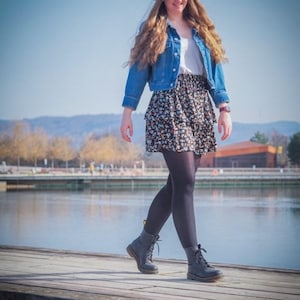 Pu&ograve; includere: Una donna indossa un cappello nero, una giacca di jeans blu, una camicia bianca, una gonna floreale, collant neri e stivali neri con lacci. Sta camminando su un molo di legno sopra un specchio d'acqua. La didascalia in fondo all'immagine recita "@schneiders.schneiderlein".