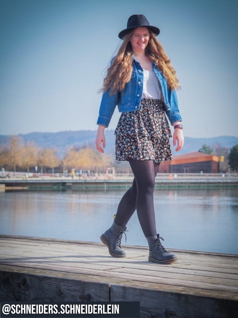 Pu&ograve; includere: Una donna indossa un cappello nero, una giacca di jeans blu, una camicia bianca, una gonna floreale, collant neri e stivali neri con lacci. Sta camminando su un molo di legno sopra un specchio d'acqua. La didascalia in fondo all'immagine recita "@schneiders.schneiderlein".