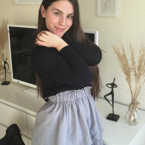 Pu&ograve; includere: Una donna indossa un maglione nero a collo alto e una gonna a strati blu chiaro. &Egrave; in piedi davanti a un muro bianco con opere d'arte e una TV.