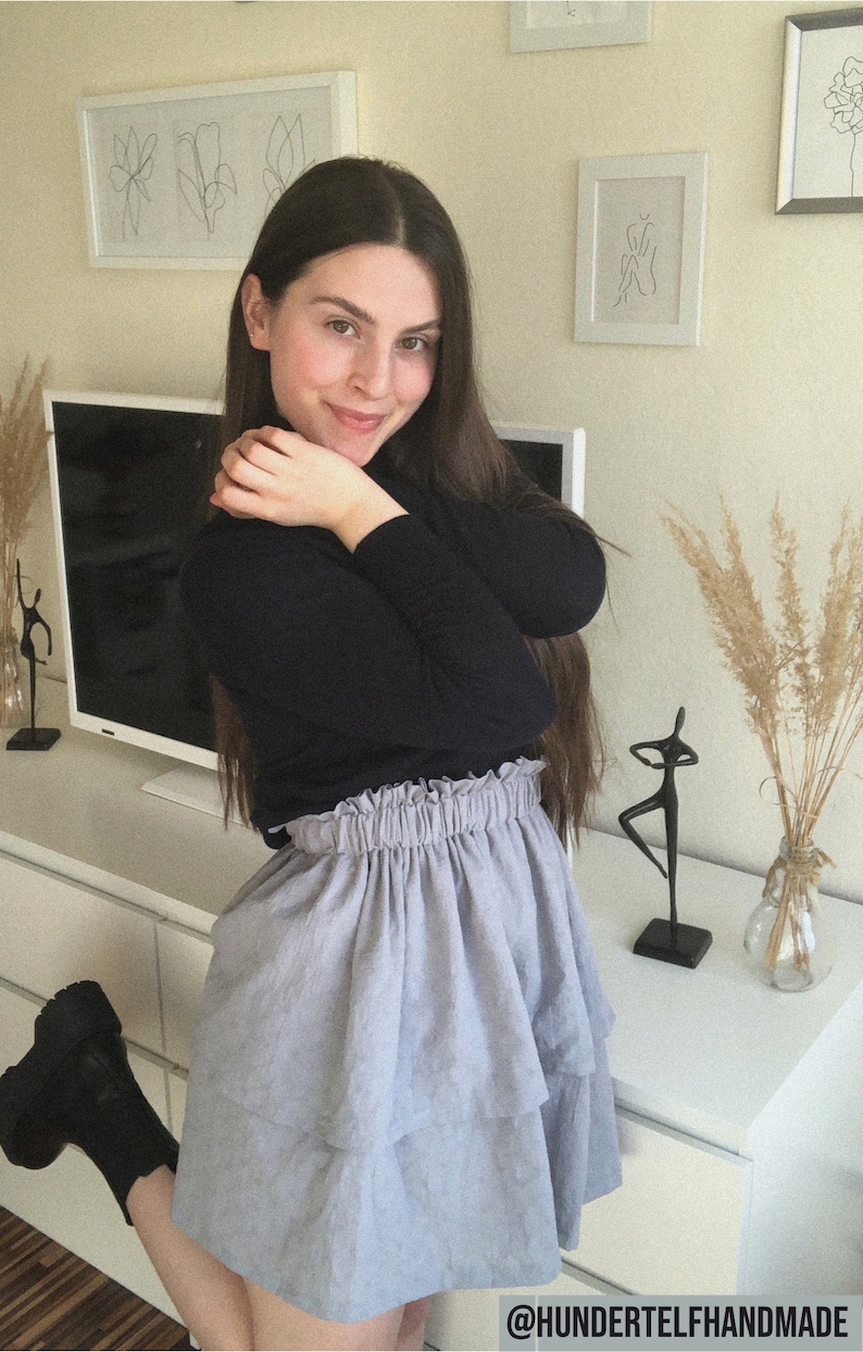 Pu&ograve; includere: Una donna indossa un maglione nero a collo alto e una gonna a strati blu chiaro. &Egrave; in piedi davanti a un muro bianco con opere d'arte e una TV.