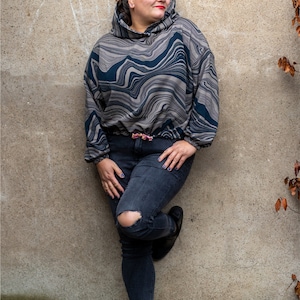 May include: A woman wearing a grey and black patterned hoodie with a drawstring. She is wearing dark blue jeans with a distressed knee and black ankle boots. The background is a grey wall with a #HOODIHEHUBERT hashtag.