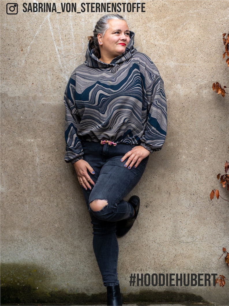 May include: A woman wearing a grey and black patterned hoodie with a drawstring. She is wearing dark blue jeans with a distressed knee and black ankle boots. The background is a grey wall with a #HOODIHEHUBERT hashtag.